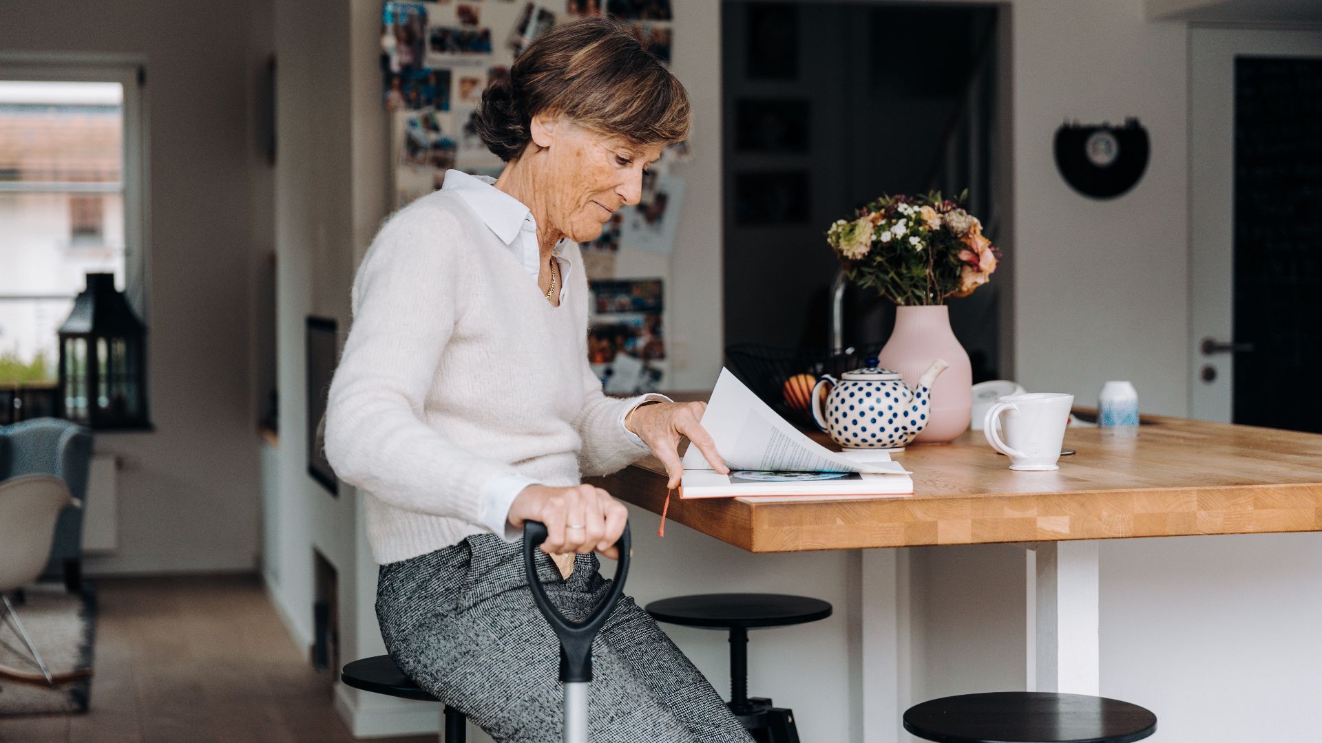 Elderly woman sitting in a cozy kitchen, reading a book. Warm and homely atmosphere with flowers and decorations.