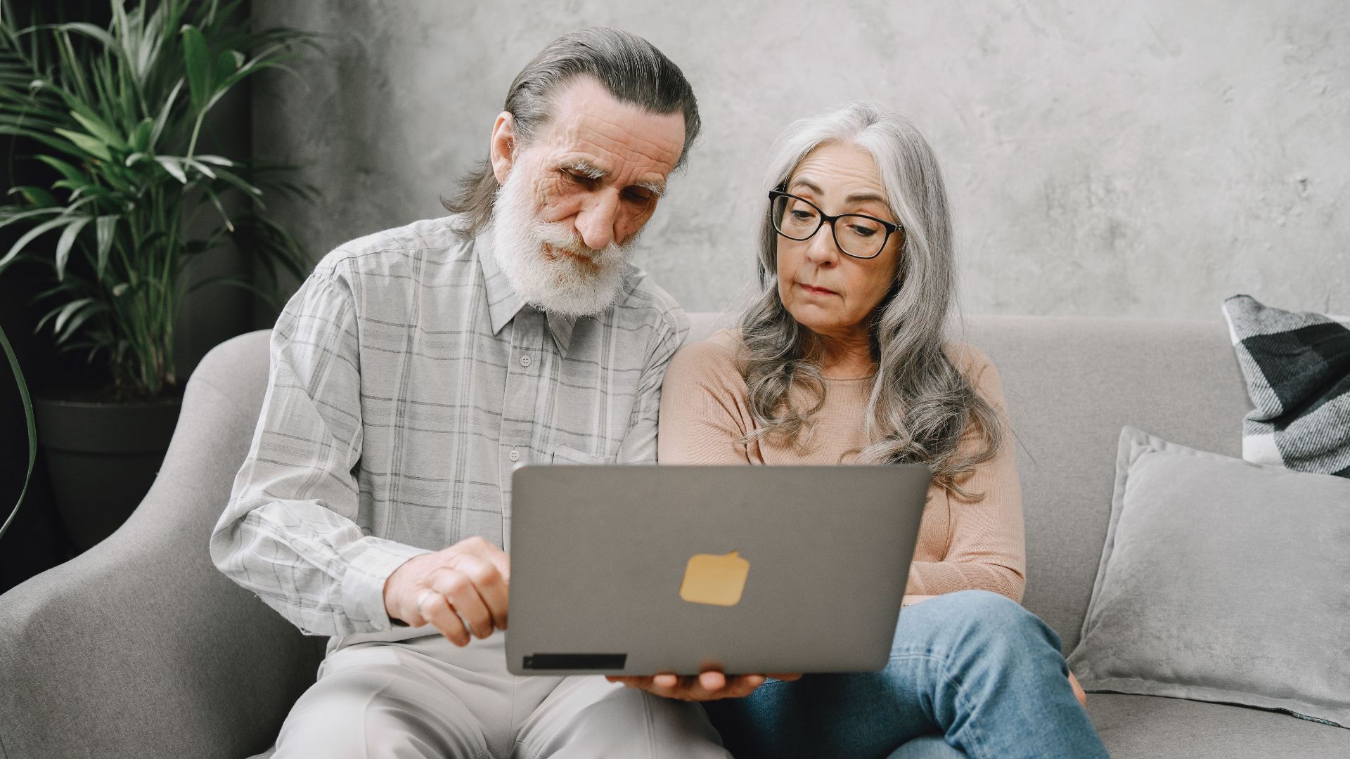 Senior couple sitting on a sofa using a laptop indoors, embracing technology together.