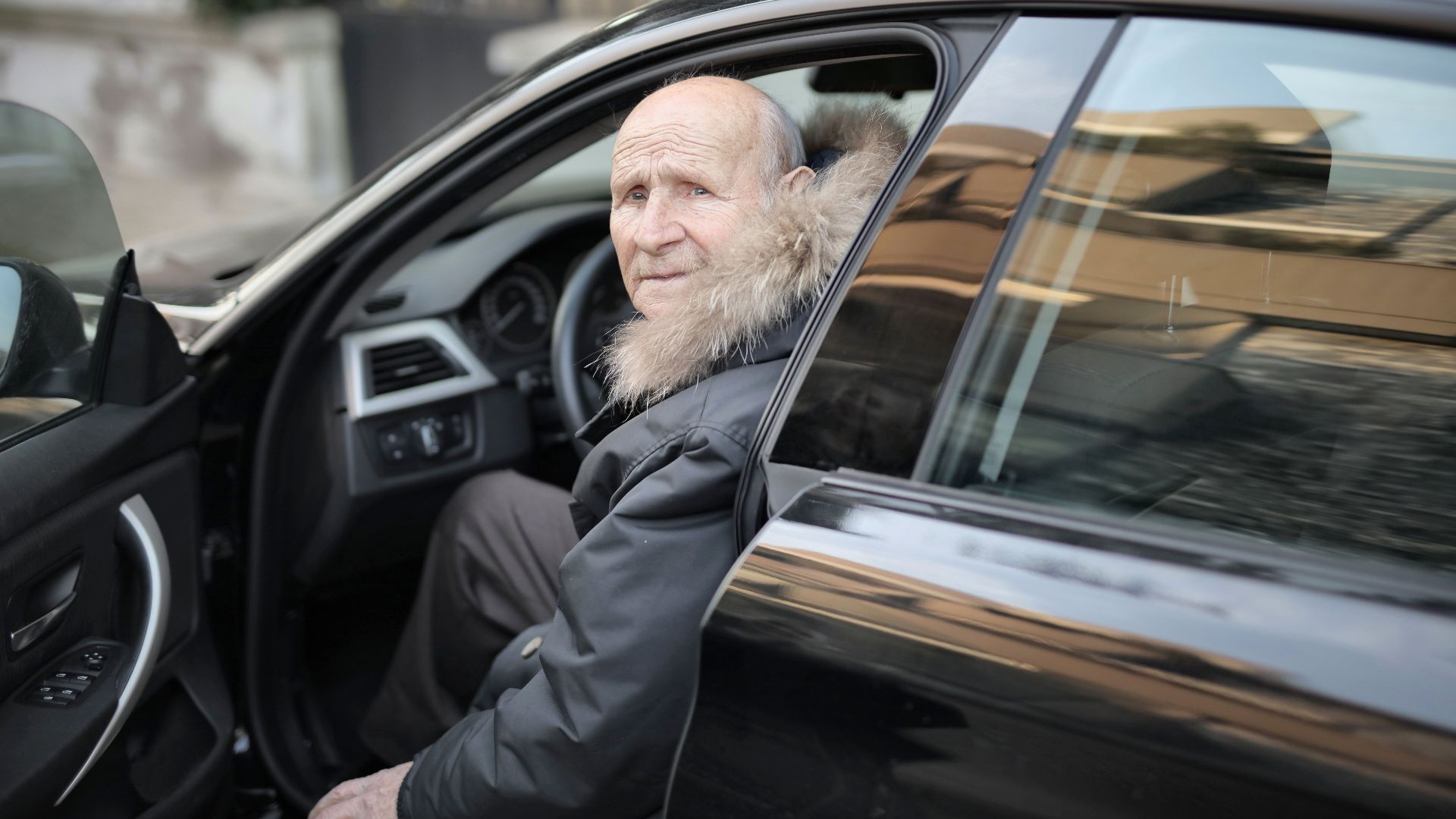 An elderly man in a winter coat stepping out of a parked car outdoors.