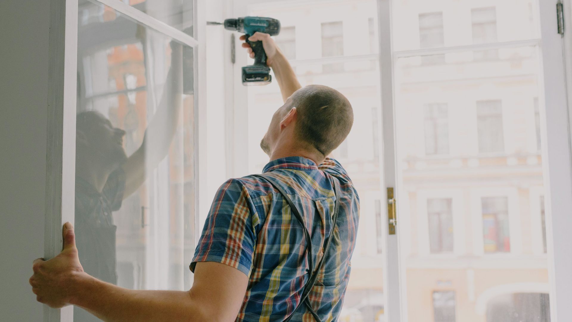 Back view of worker in checkered shirt drilling window frame during renovation process in apartment