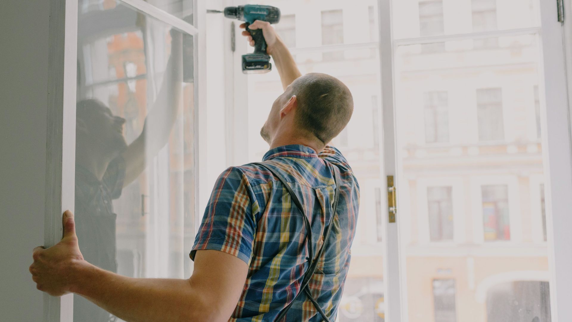 Back view of worker in checkered shirt drilling window frame during renovation process in apartment