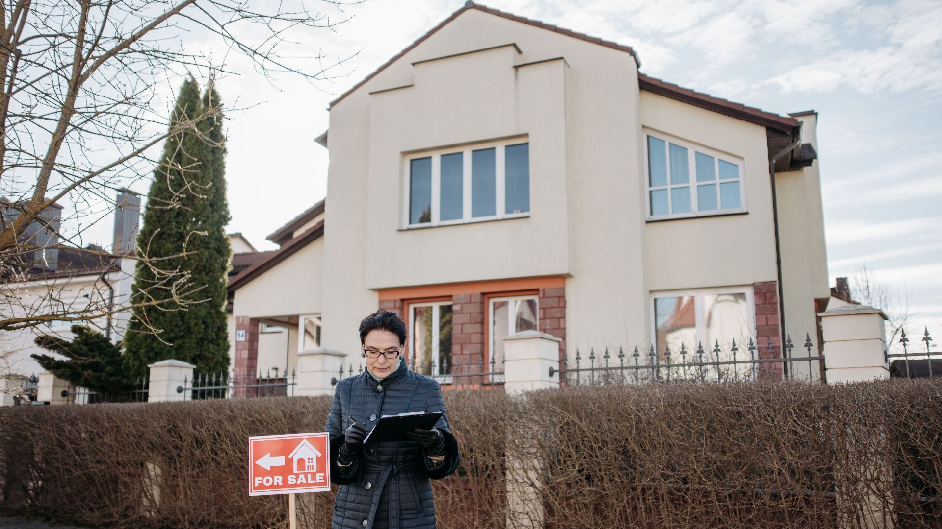 A real estate agent standing outside a house with a 'For Sale' sign, ready for viewing.