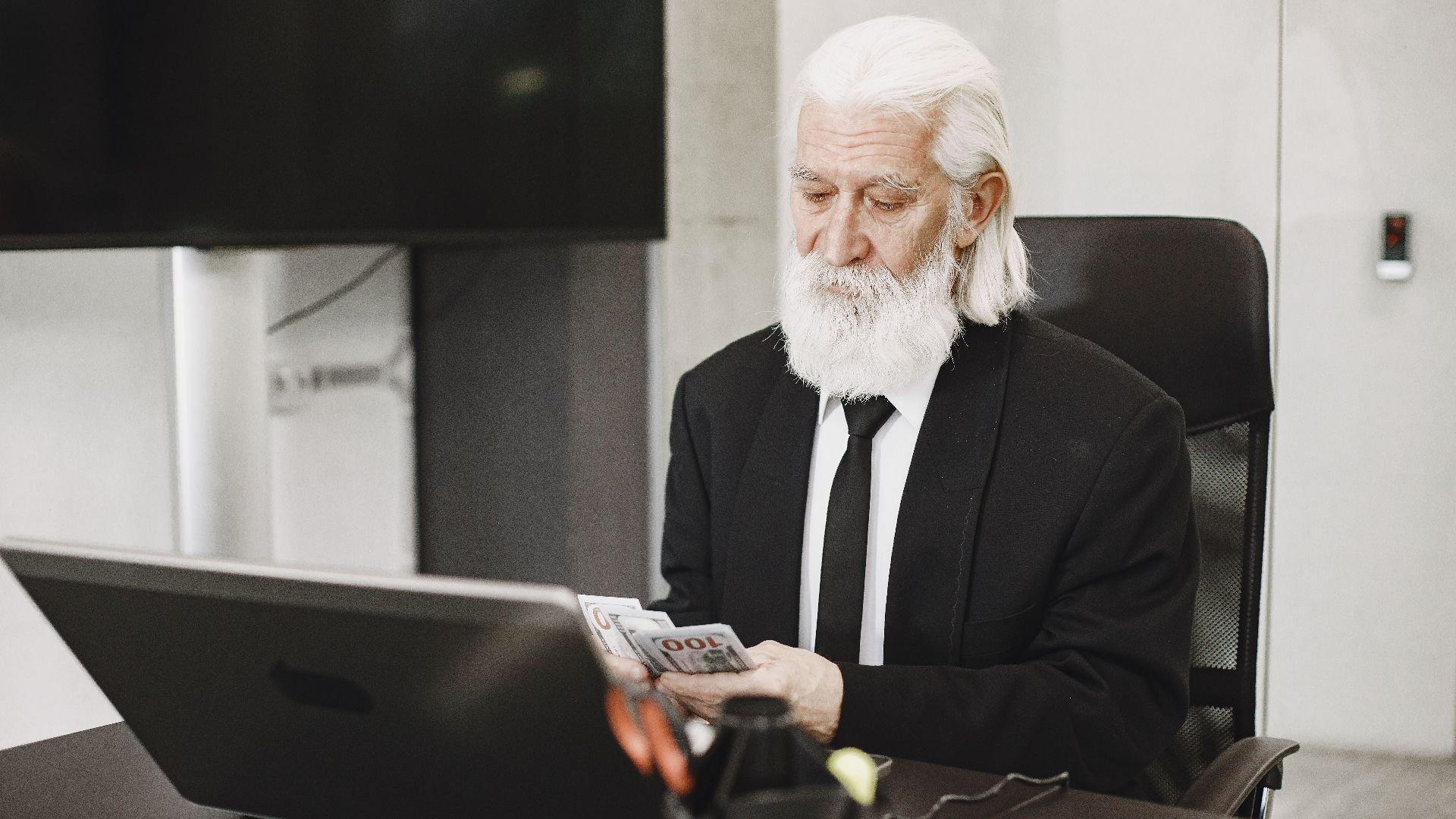 White-haired man in suit counting money at a modern office desk with laptop.