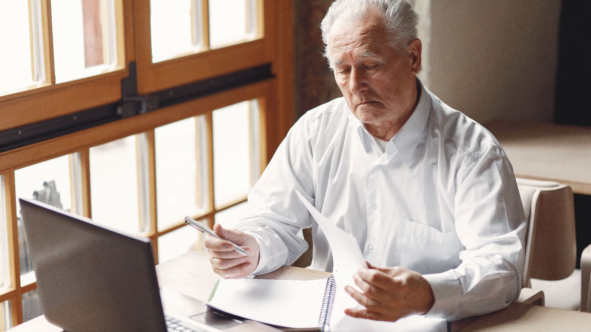 Serious gray haired aged man in white shirt sitting at table and browsing laptop while checking notes in modern office