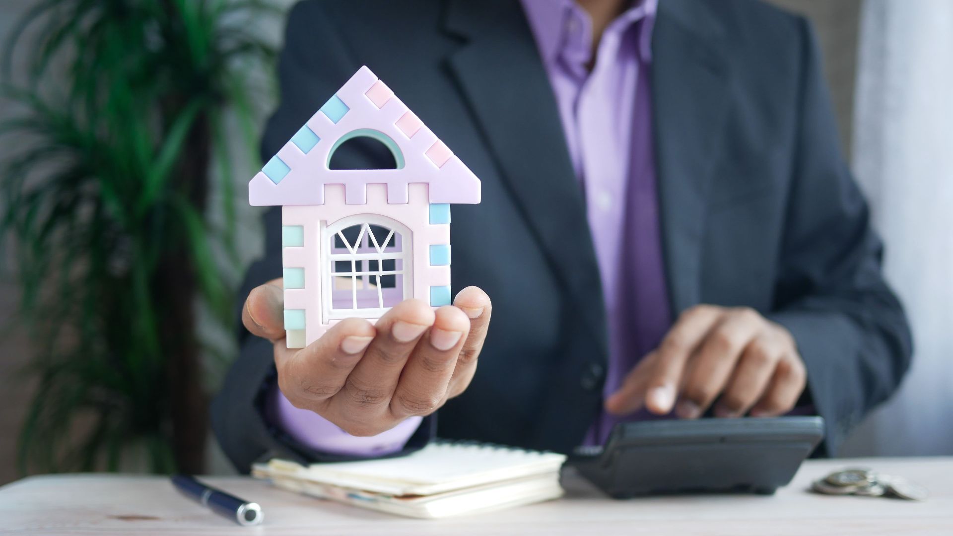 Businessman in suit holds model house, calculates real estate investments.