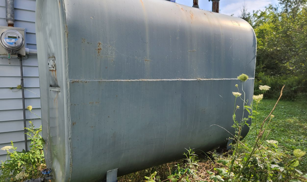 An oil tank hidden on the side of a suburban house. The tank is painted gray and nestled next to vegetation.