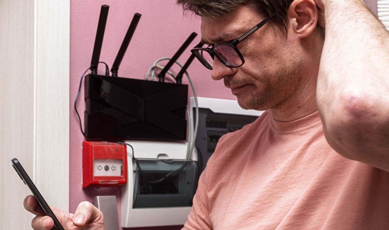 Concerned man checks smartphone near WIFI router and switchboard during home internet problem, confusion and weak connection