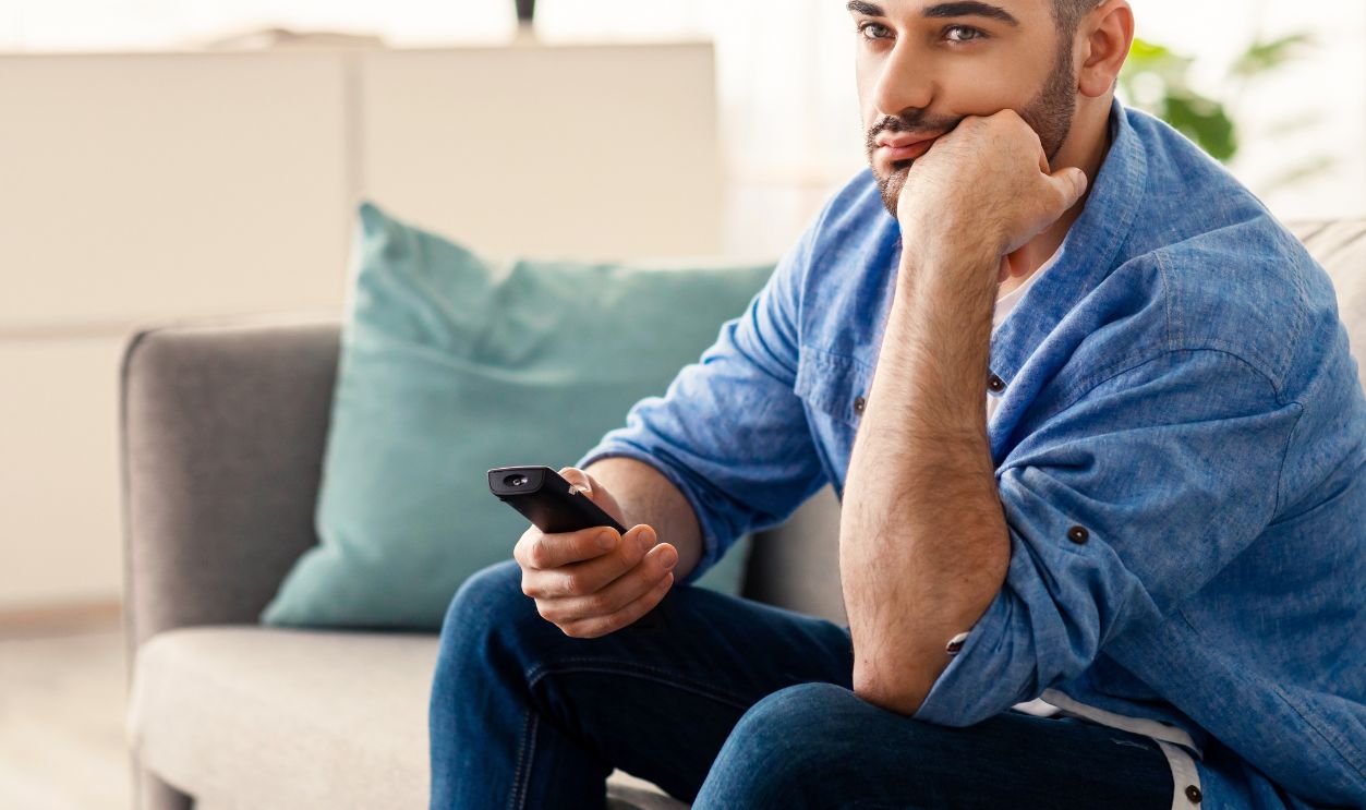 Boring Television Program. Bored Jewish Man Watching TV Switching Channels With Remote Control Sitting On Sofa At Home. Discontented Viewer Watching Uninteresting Movie, Feeling Lonely