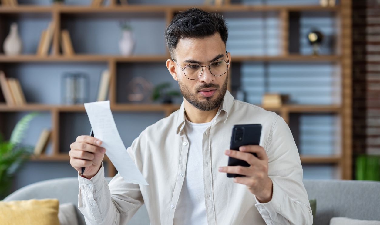 young man sitting on sofa at home, holding letter, receipt and worriedly looking at mobile phone, checking bill.