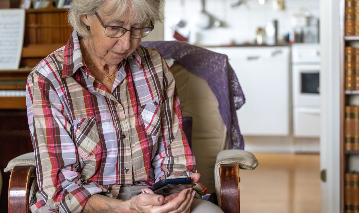 An elderly, gray-haired woman sits thoughtfully looking down at the screen of her mobile phone.