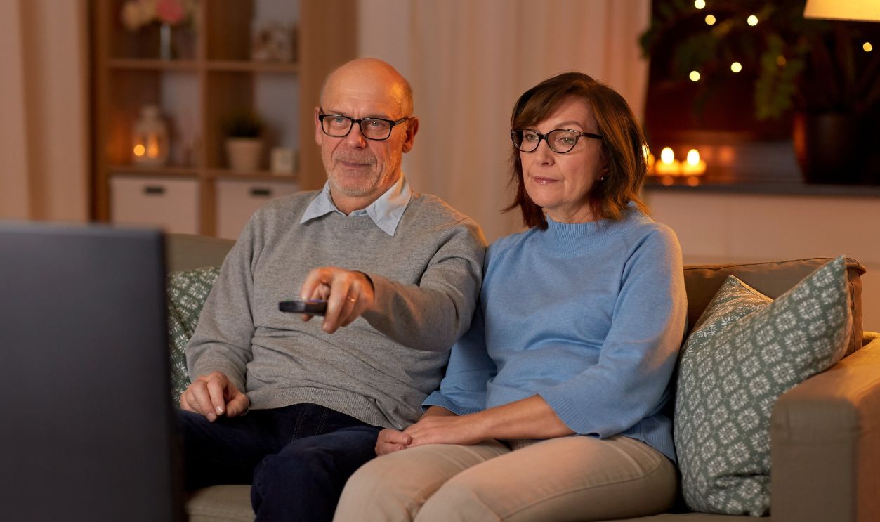 holidays, drinks and people concept - senior couple with remote control watching tv at home in evening