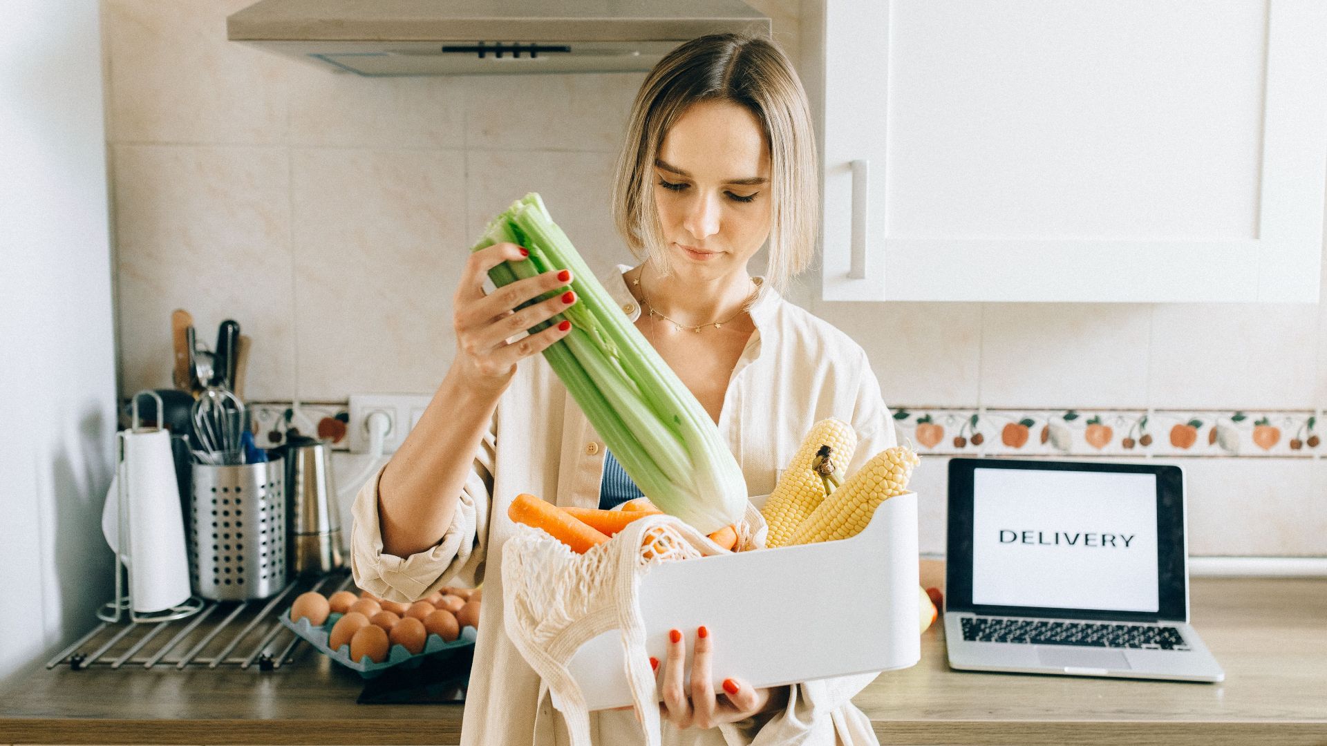Woman unpacks fresh vegetables from delivery box in a modern kitchen setup with a laptop nearby.