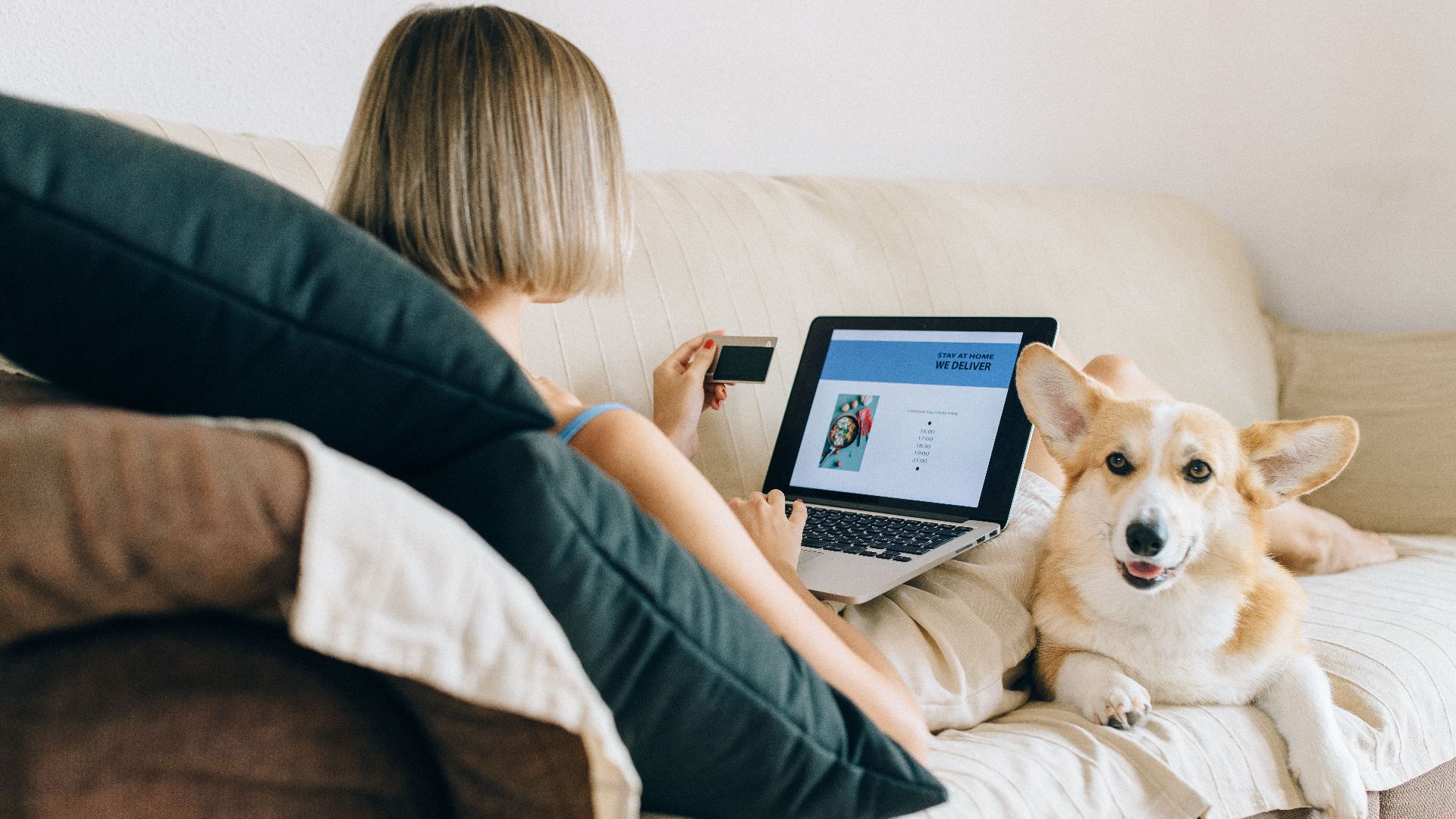 A woman using a laptop for online shopping with her corgi dog beside her on the couch.