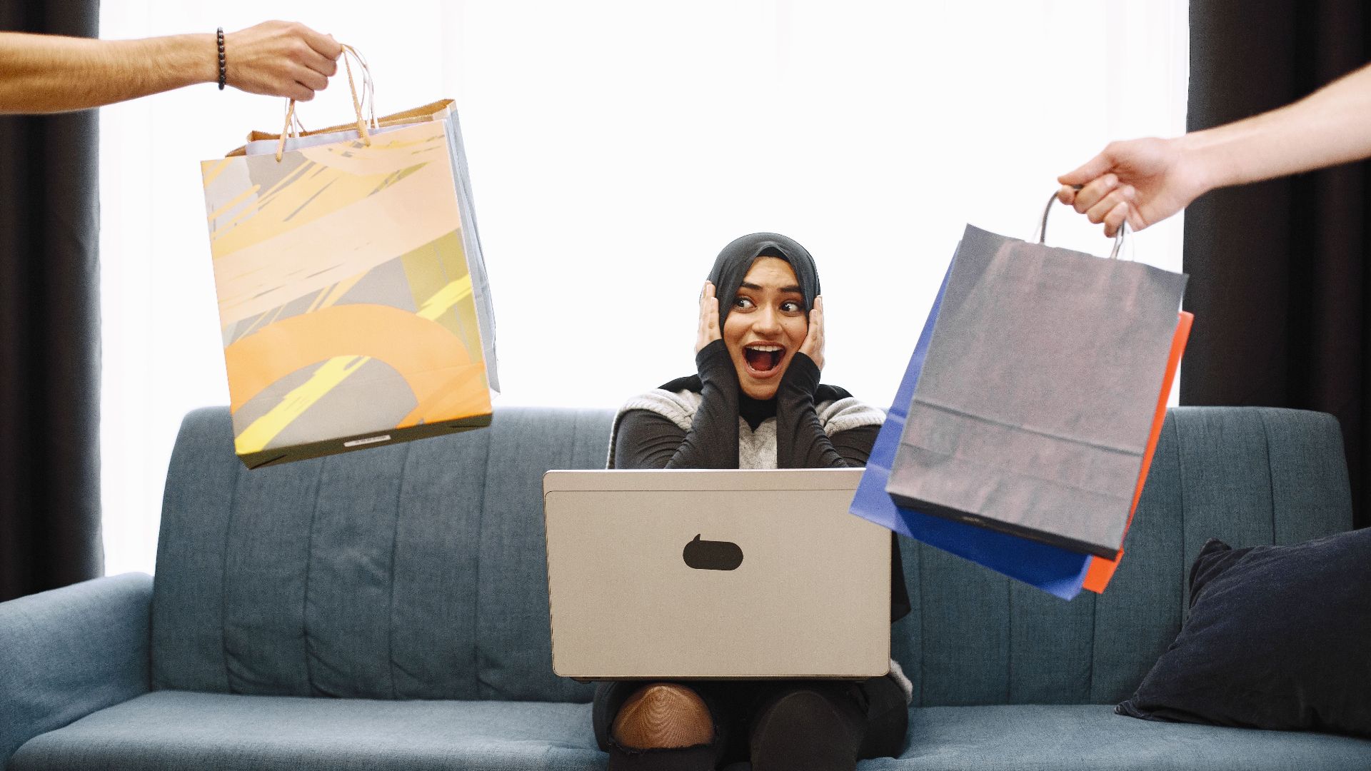 A woman in a hijab excitedly shopping online, receiving bags while sitting on a couch with a laptop.