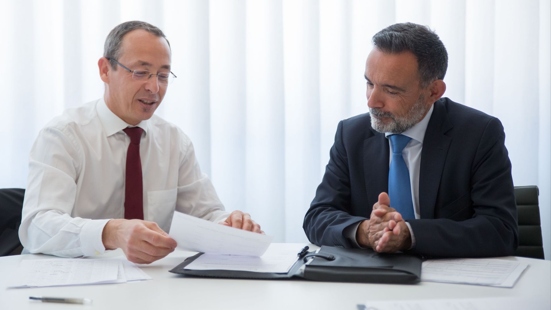 Two businessmen in suits having a discussion over documents in a bright office setting.
