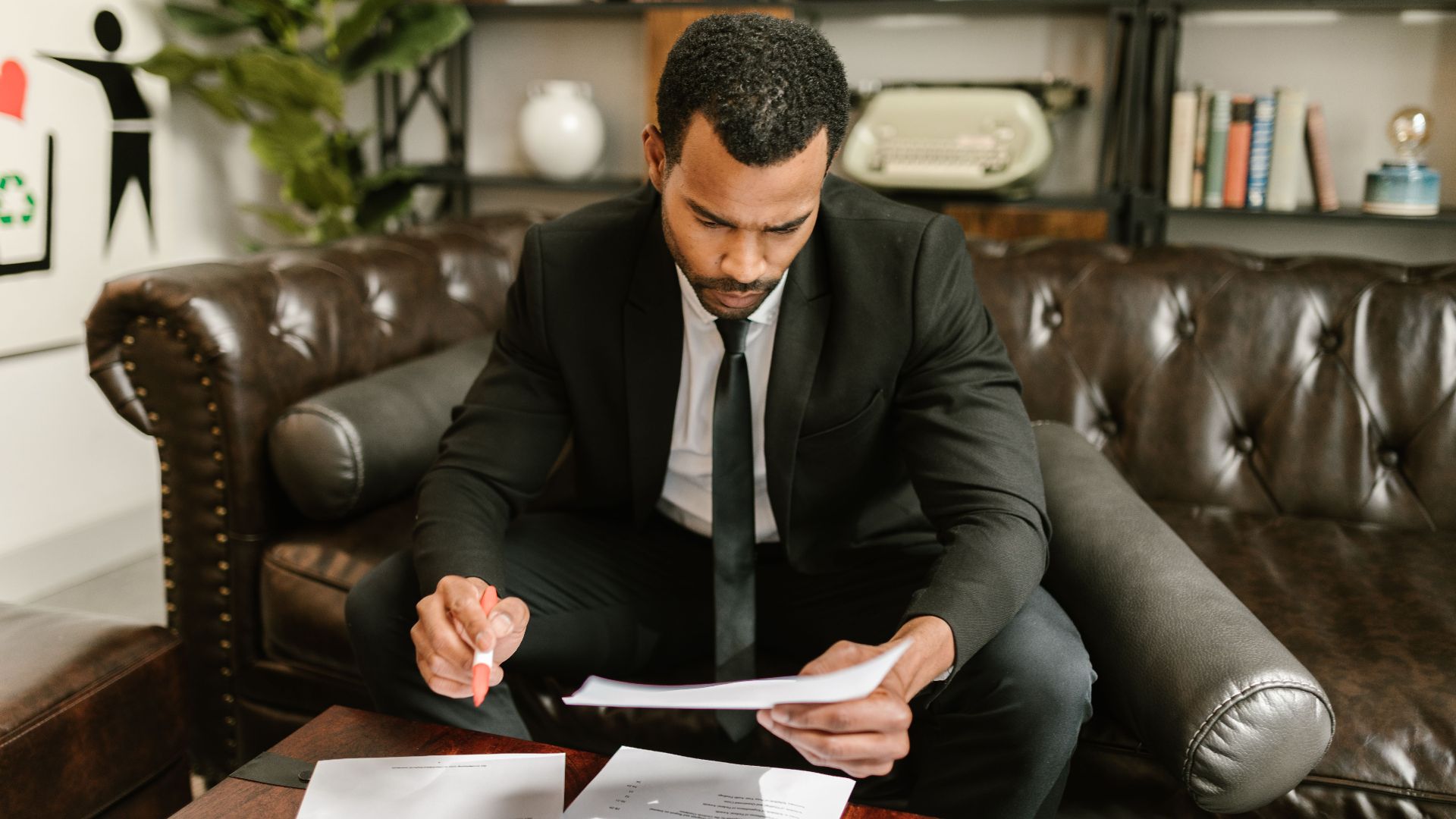 A businessman in a suit attentively reviews paperwork on a brown leather sofa.