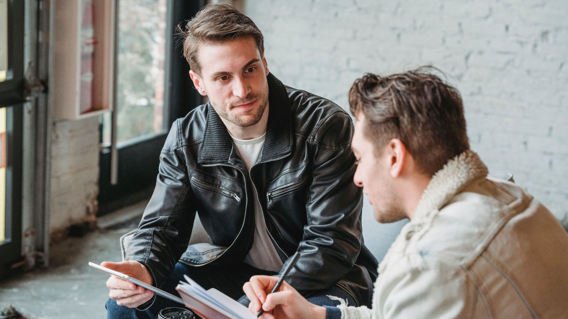 Concentrated young man writing in notebook while doing project with colleague sitting nearby with tablet