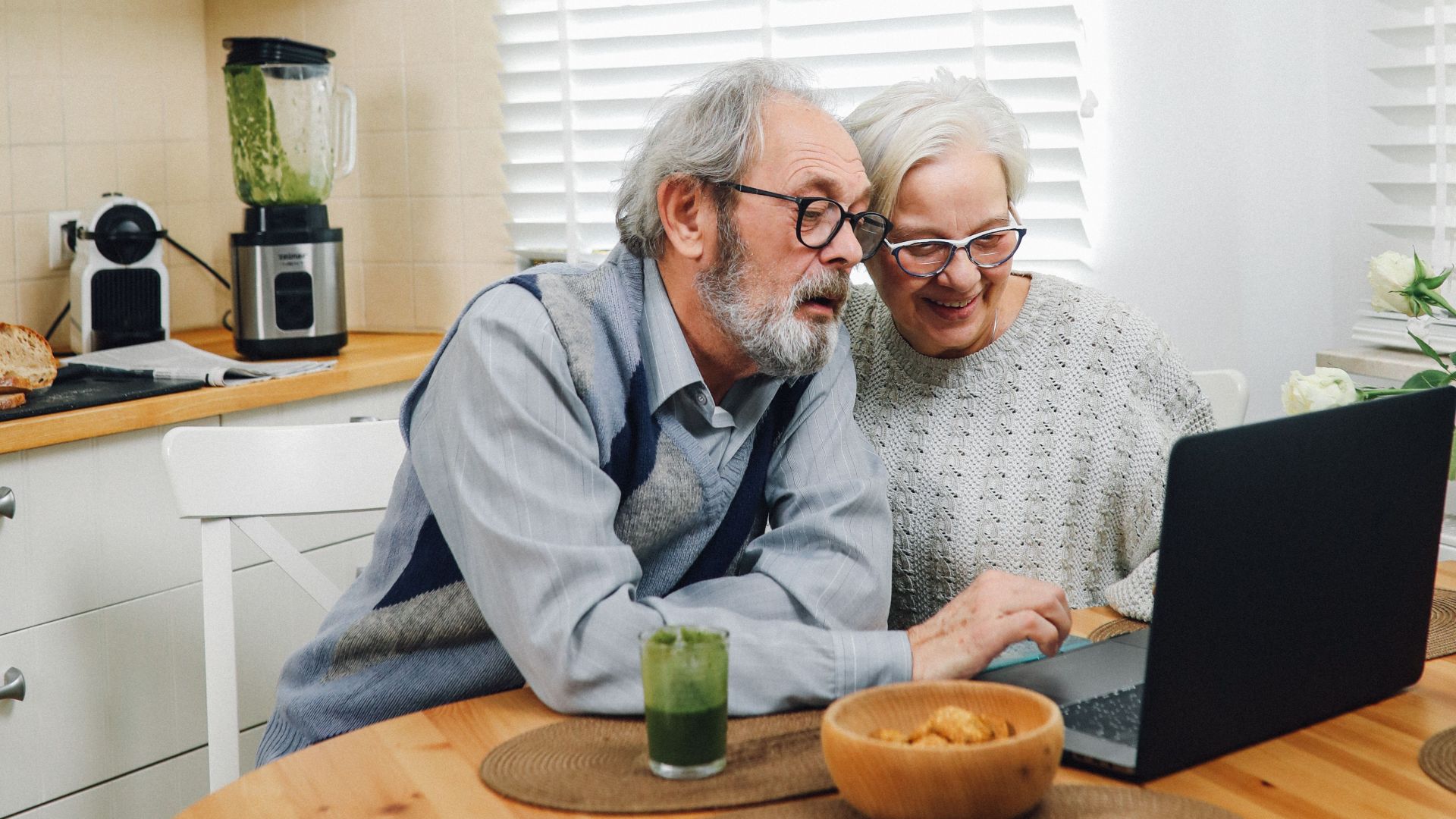 Elderly couple using a laptop in a bright kitchen, sharing moments of happiness and togetherness.