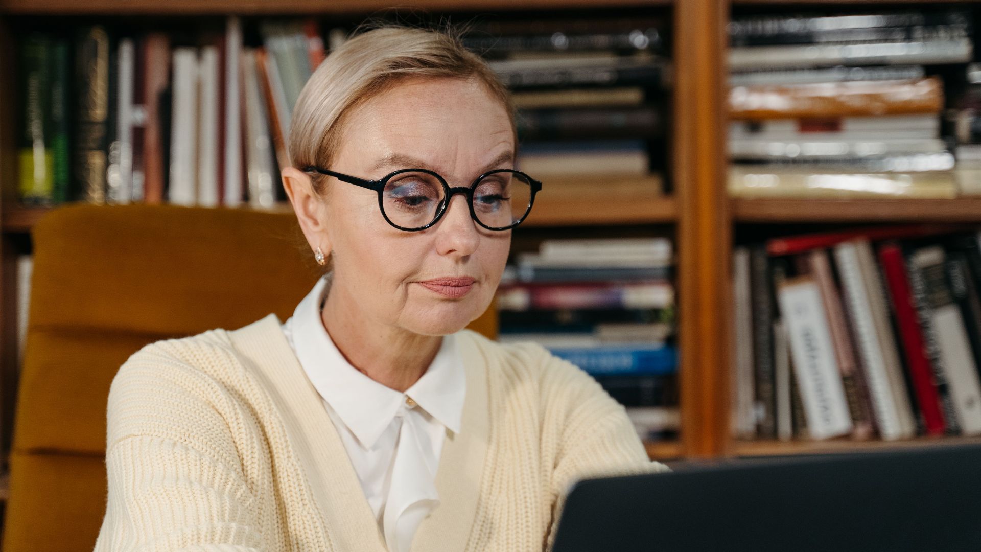 A senior woman wearing glasses focusing on her laptop in a cozy library environment.