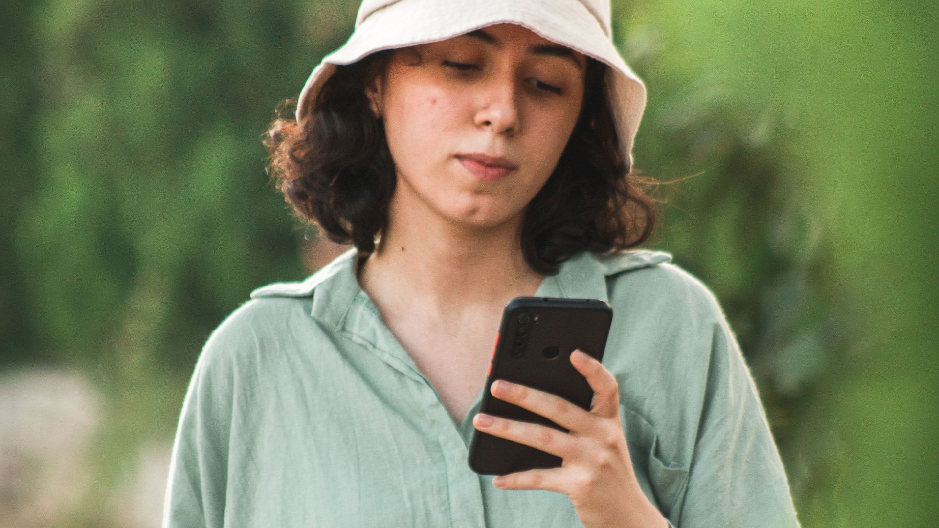 A young woman in a bucket hat uses her smartphone while walking outdoors.