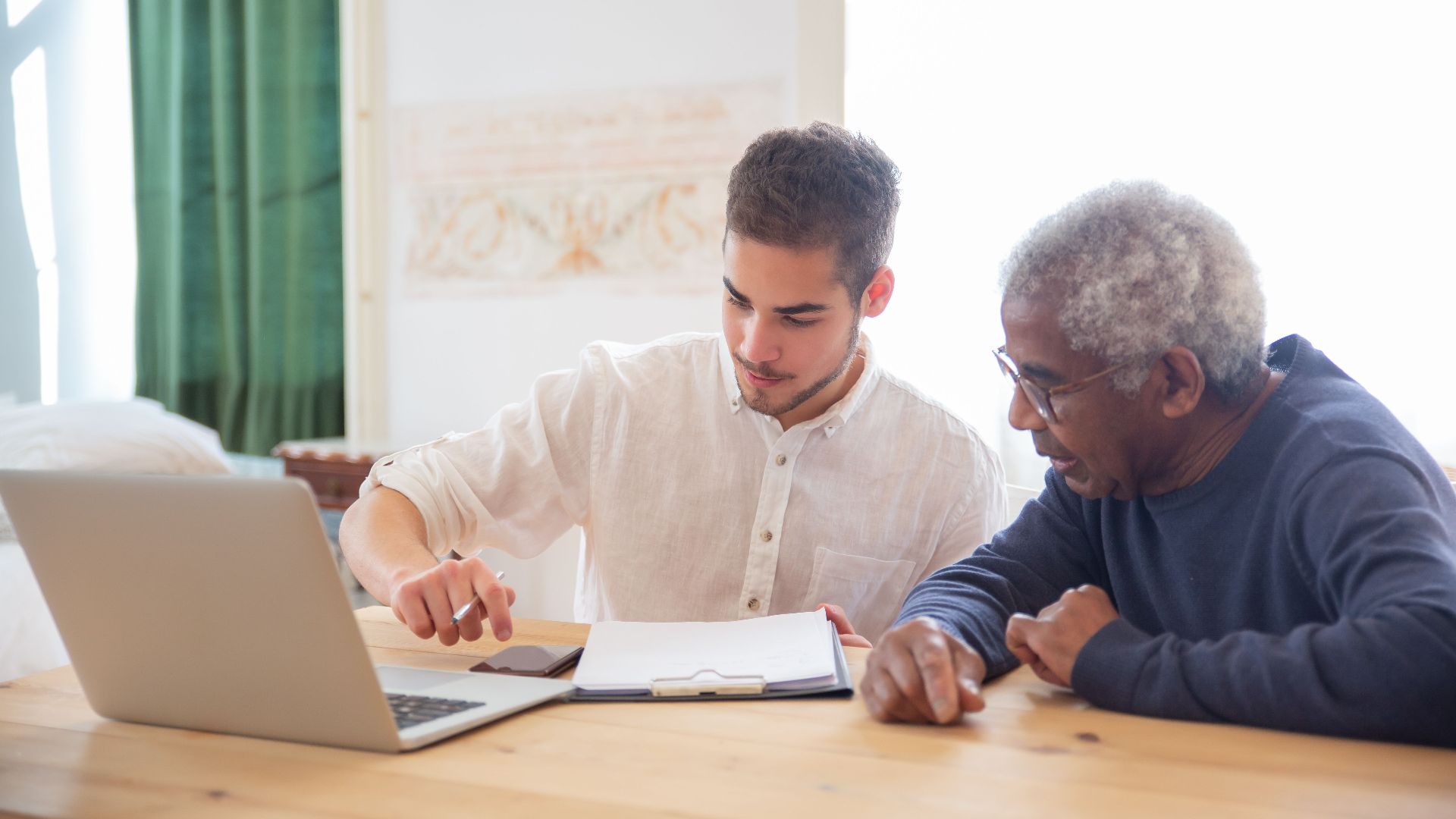 Young man teaches elderly gentleman to use technology at home.