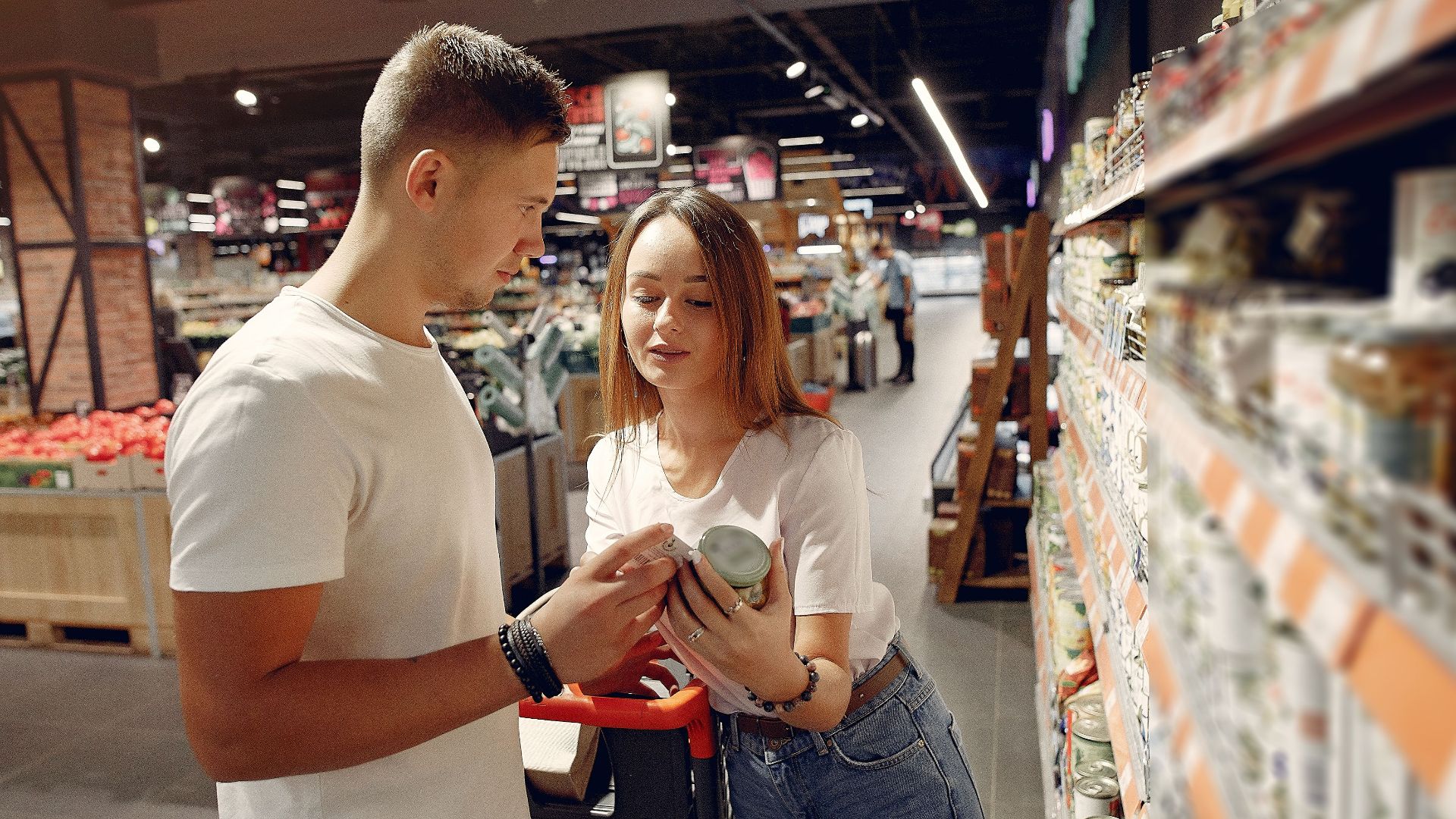 From above of young woman in casual wear showing canned food for man while standing with grocery trolley near shelves with products in modern supermarket