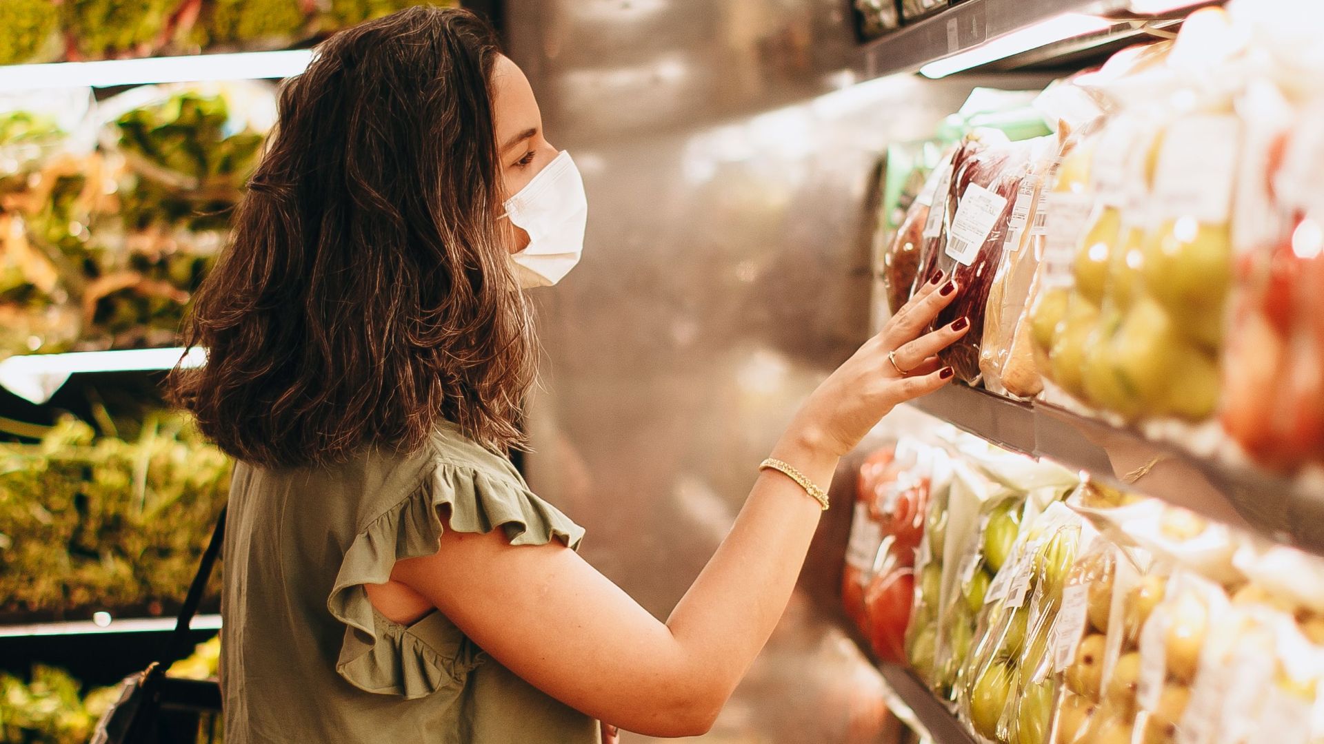 Woman wearing a mask choosing fresh produce in a supermarket aisle.