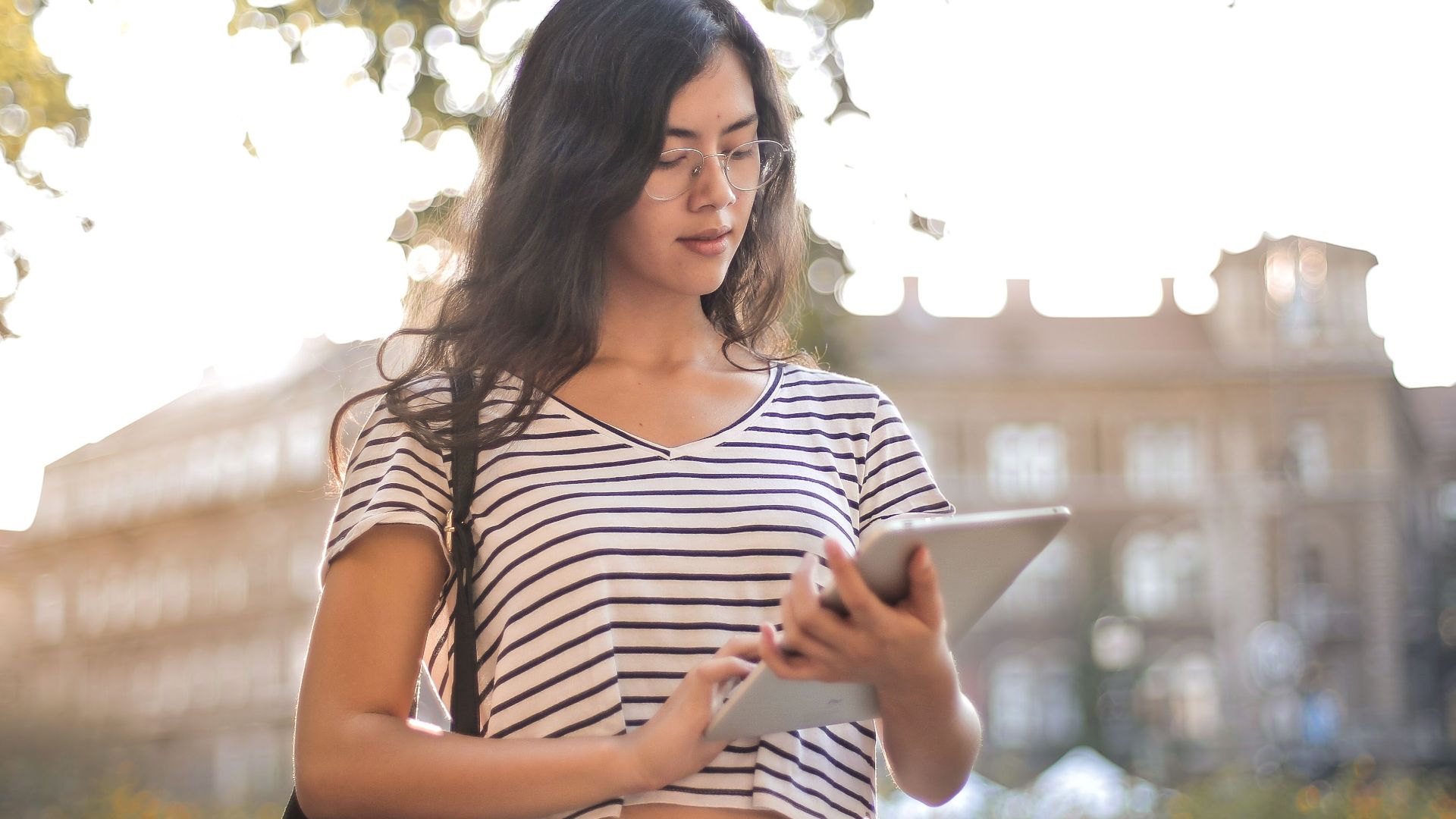 A young woman in a striped shirt and ripped jeans using a tablet in a sunny park.