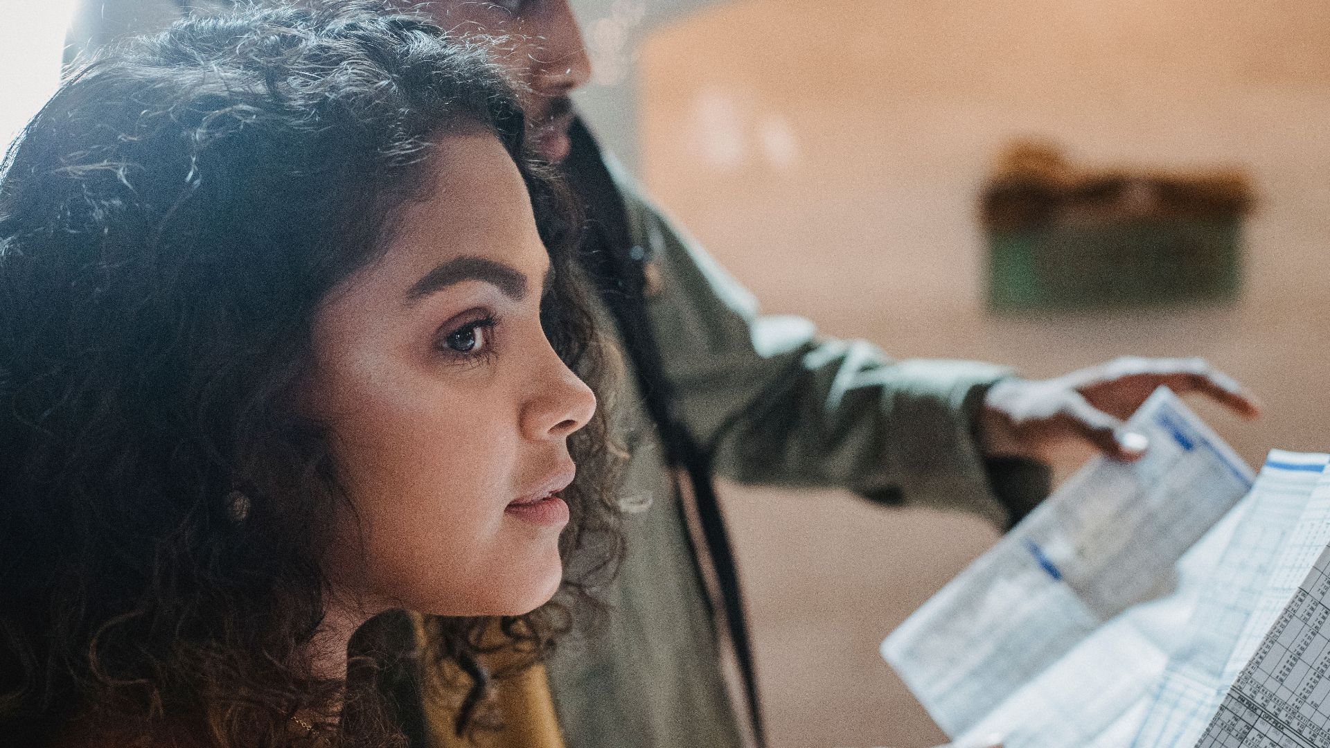 Side view of thoughtful Hispanic woman with dark curly hair standing with black friend reading long paper with tables