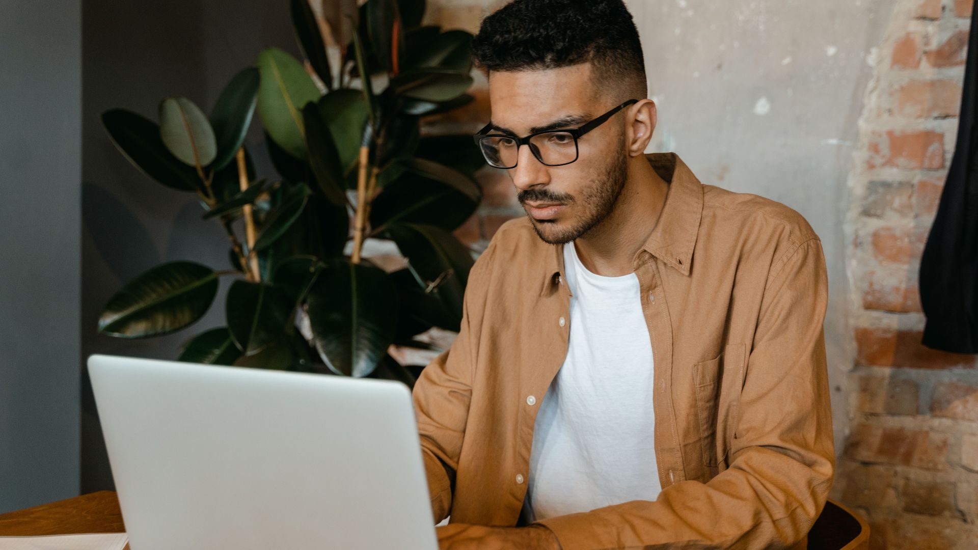 A man in glasses working on a laptop in a cozy cafe with brick walls and plants.