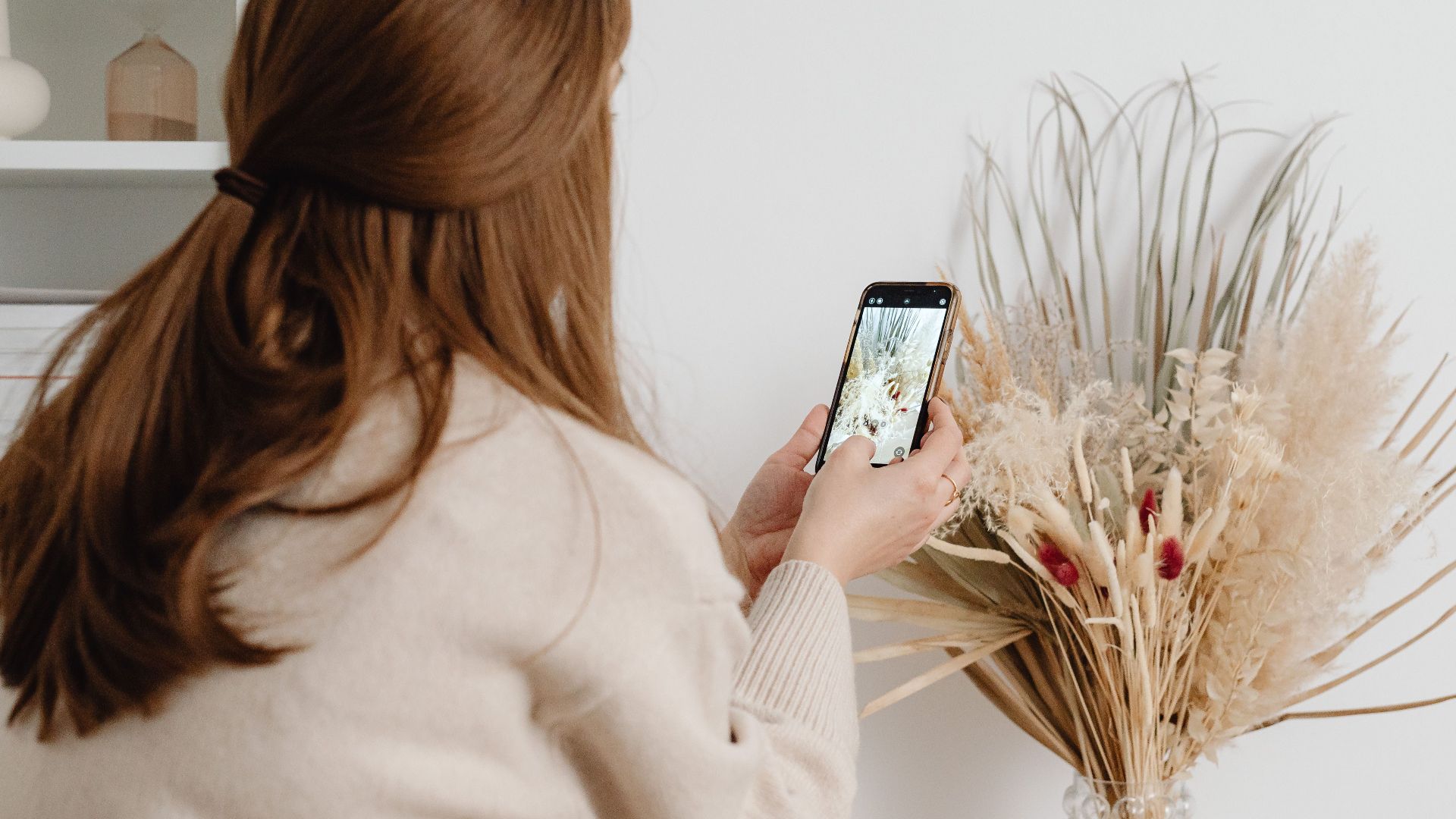 A woman takes a picture of a dried floral arrangement in a stylish home setting.