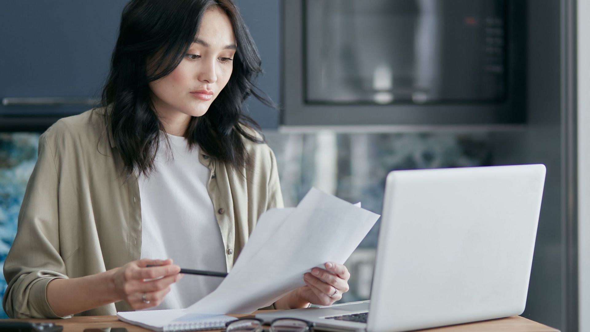Concentrated woman reviewing documents and working on a laptop at home office setup in kitchen.