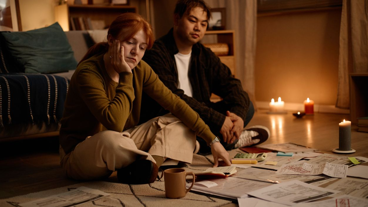 Shutterstock-2809299903, Young adult couple reviewing bills and household paperwork while sitting on floor, showing financial stress and budgeting challenges. Useful for debt, inflation, personal finance marketing