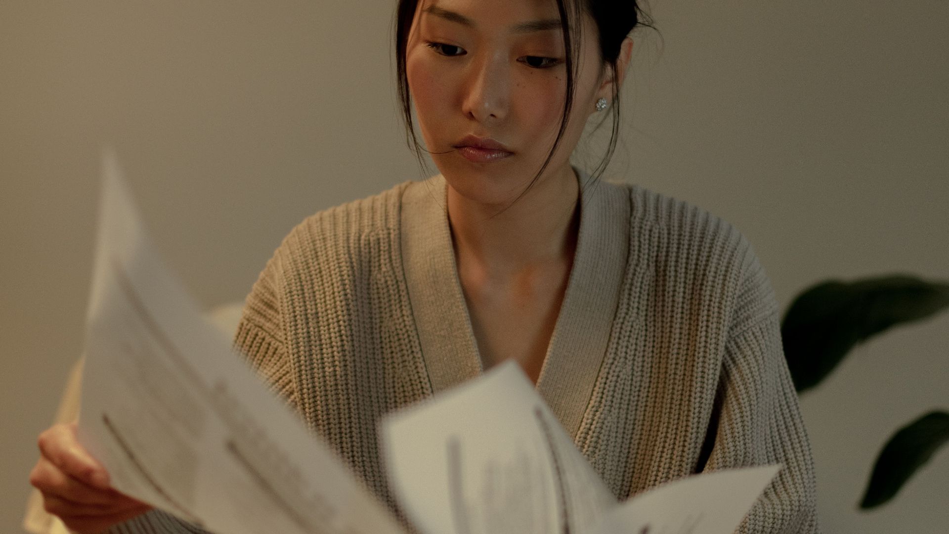 A woman with long black hair sits indoors, reading papers in a cozy and warm setting.