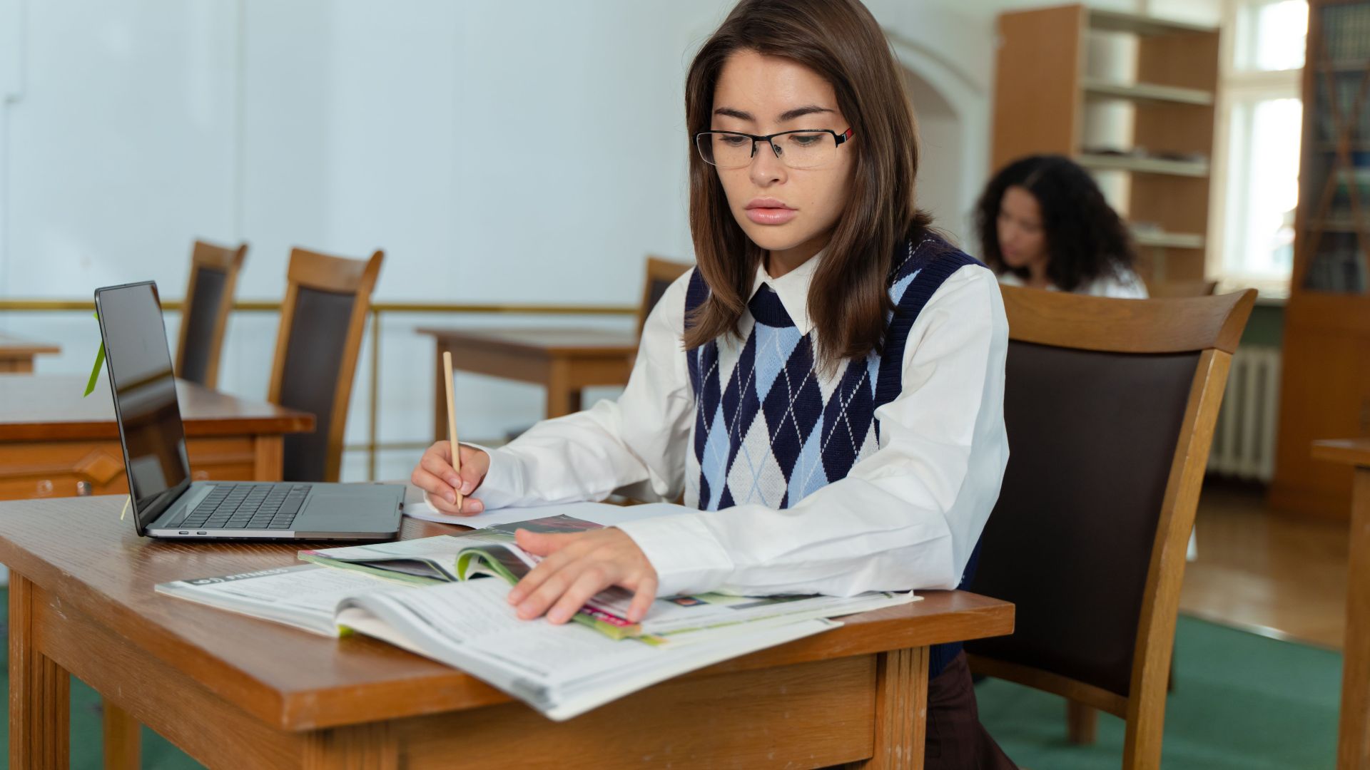 A focused young woman studying with textbooks and a laptop in a quiet library setting.