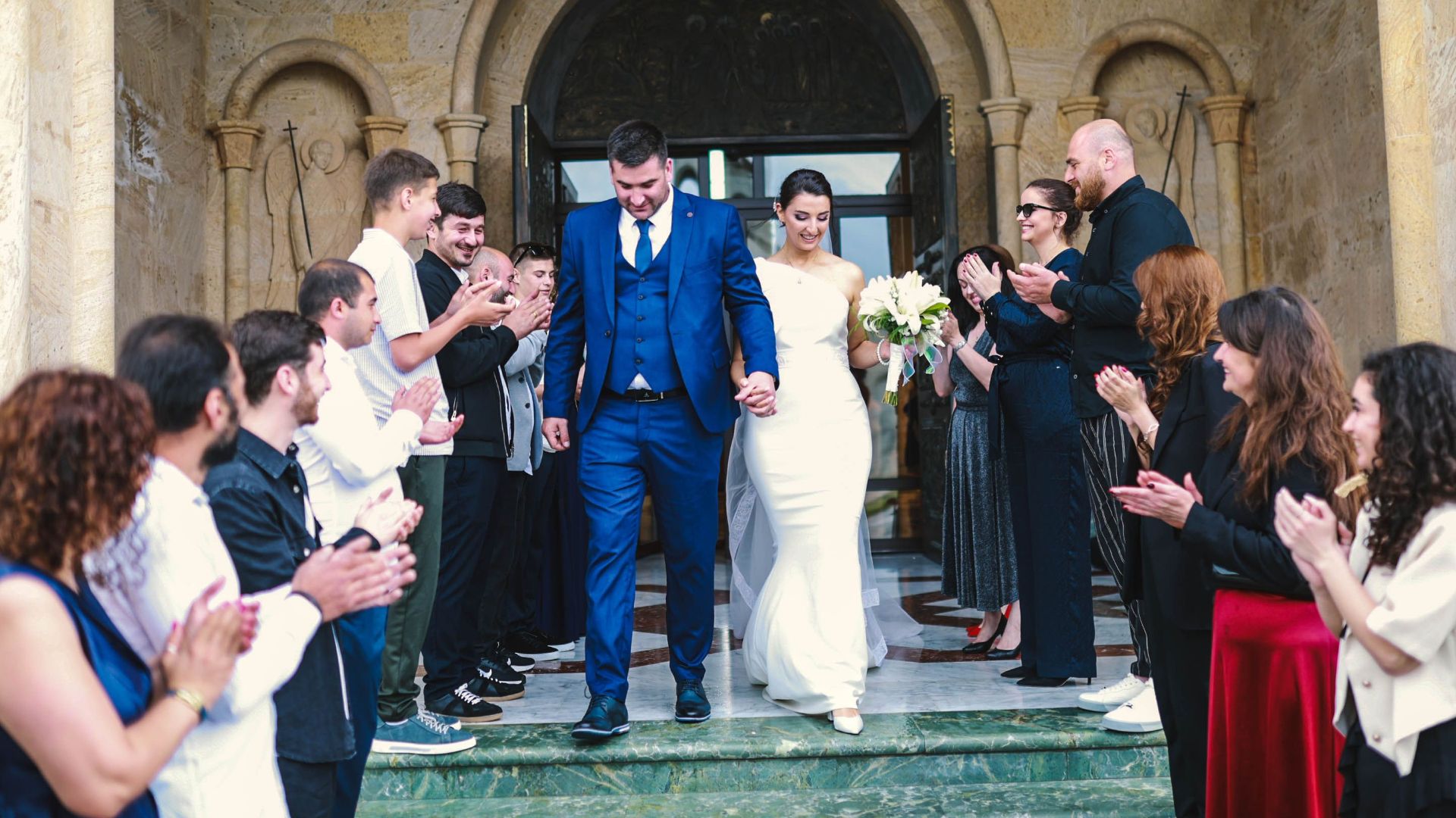 Bride and groom exiting a church, surrounded by applauding guests in an outdoor wedding ceremony.