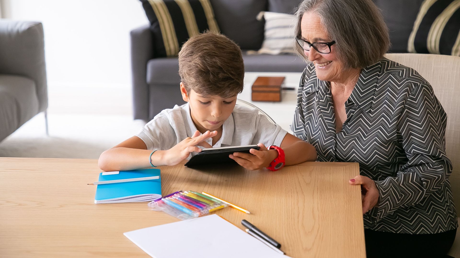 A senior woman smiles while helping her grandson with a tablet in a cozy living room.