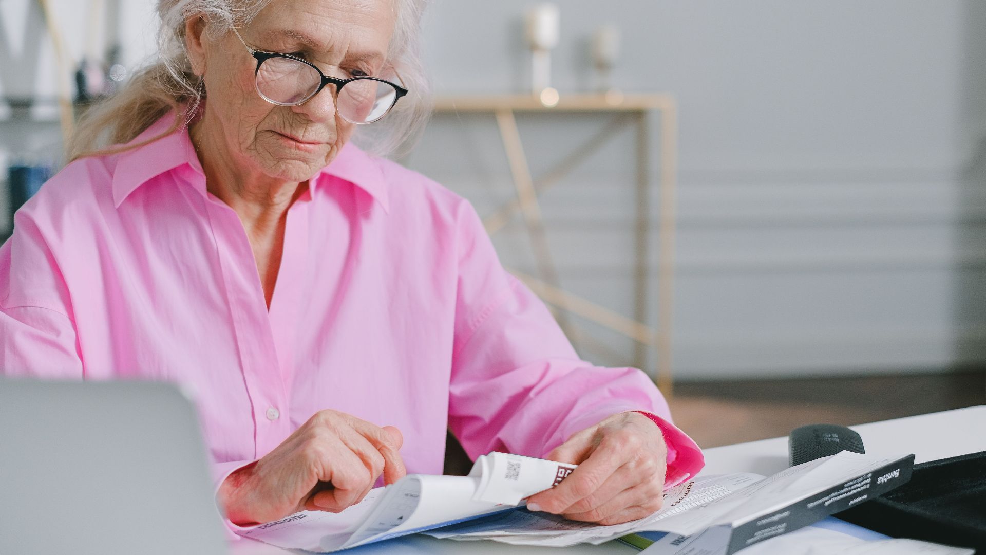 Elderly woman in pink shirt reading documents at a desk indoors.