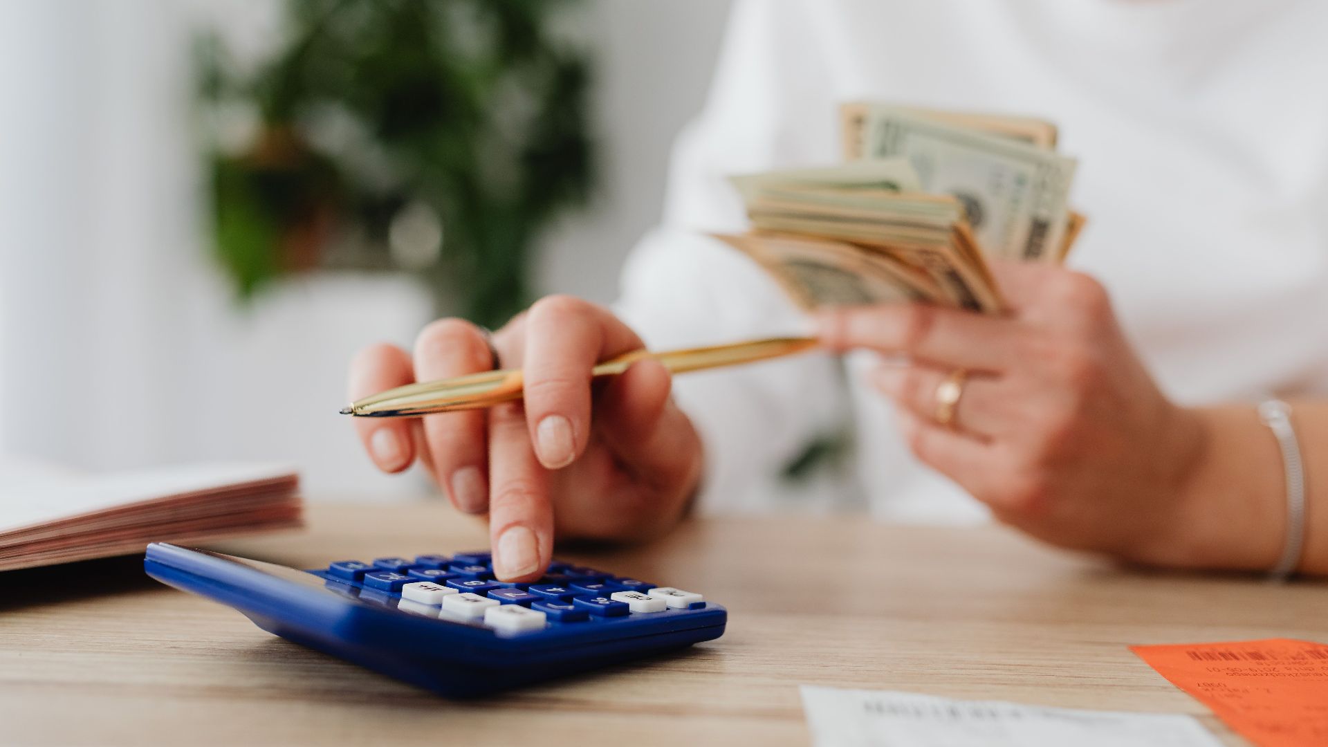 A person sits at a desk calculating finances using a calculator and holding cash.