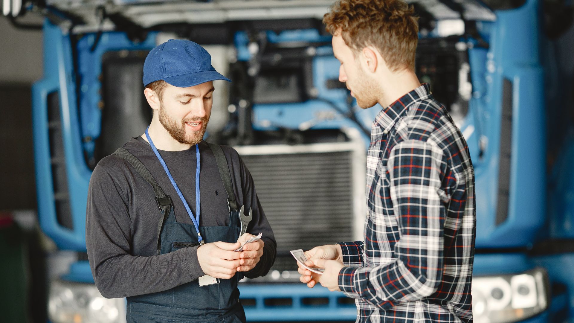 Auto mechanic in blue uniform receives payment from customer in a garage setting with a truck in the background.