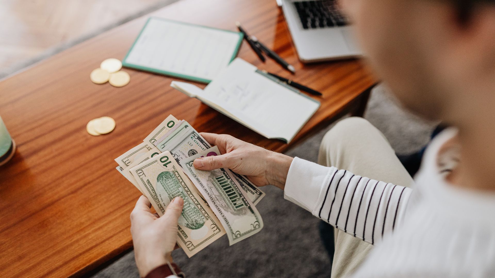 A person counts US dollar bills with a notebook and coins on the desk.
