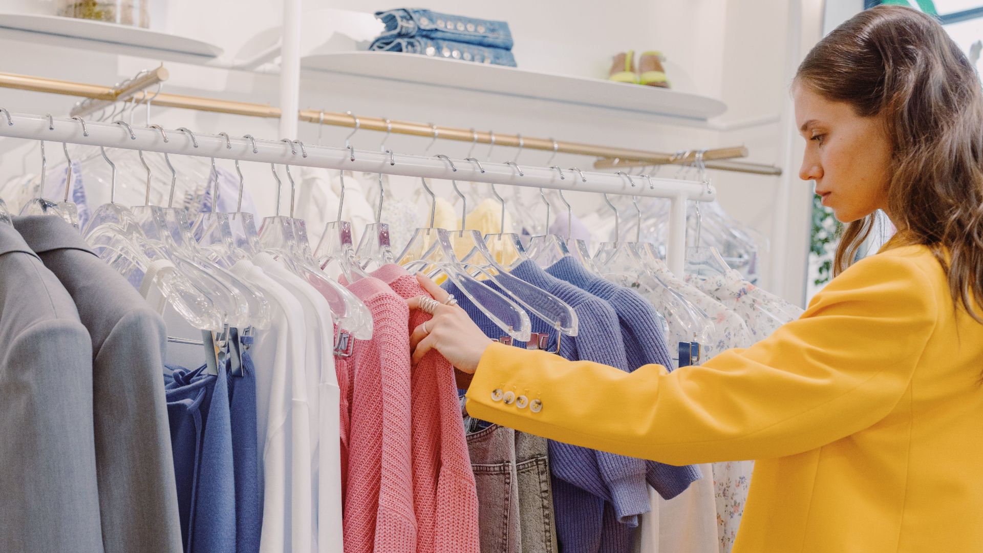 Woman browsing clothes on a rack in a clothing store, wearing a stylish yellow blazer.