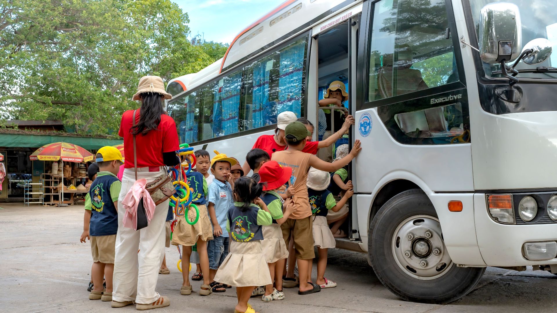Group of children and adults boarding a bus, preparing for a school trip in a sunny outdoor setting.