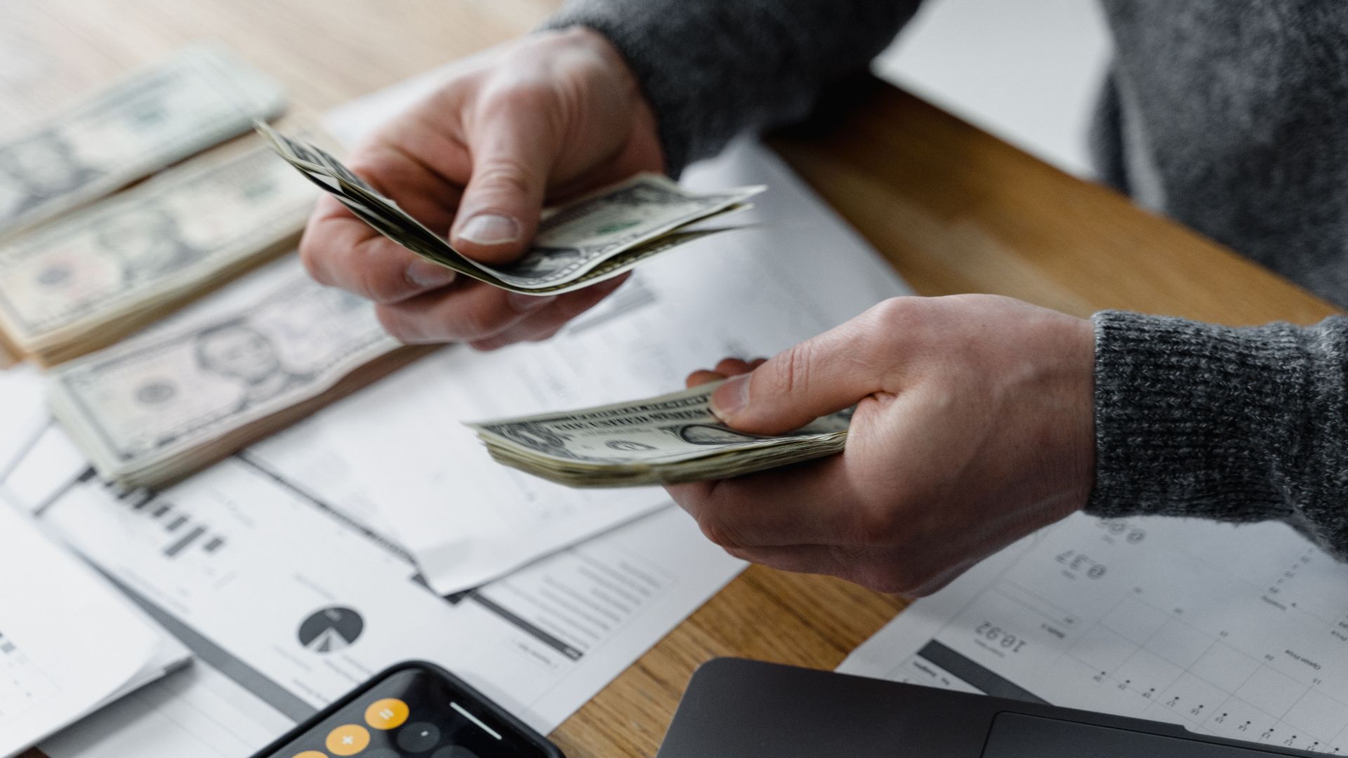 Person counting dollar bills on a desk with financial documents and a calculator in the background.