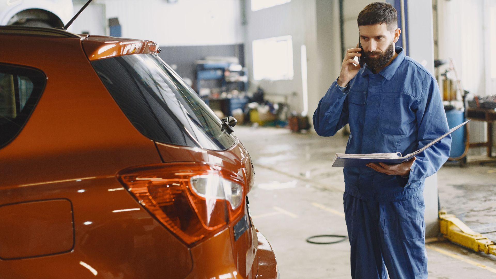 Mechanic in a blue uniform talks on phone beside a red car in an auto repair shop.