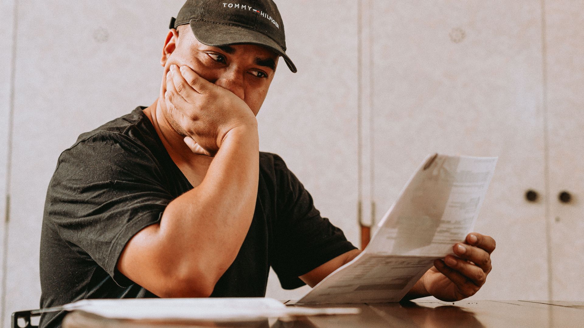 A man sits indoors, deeply focused on reviewing paperwork, expressing concern and contemplation.