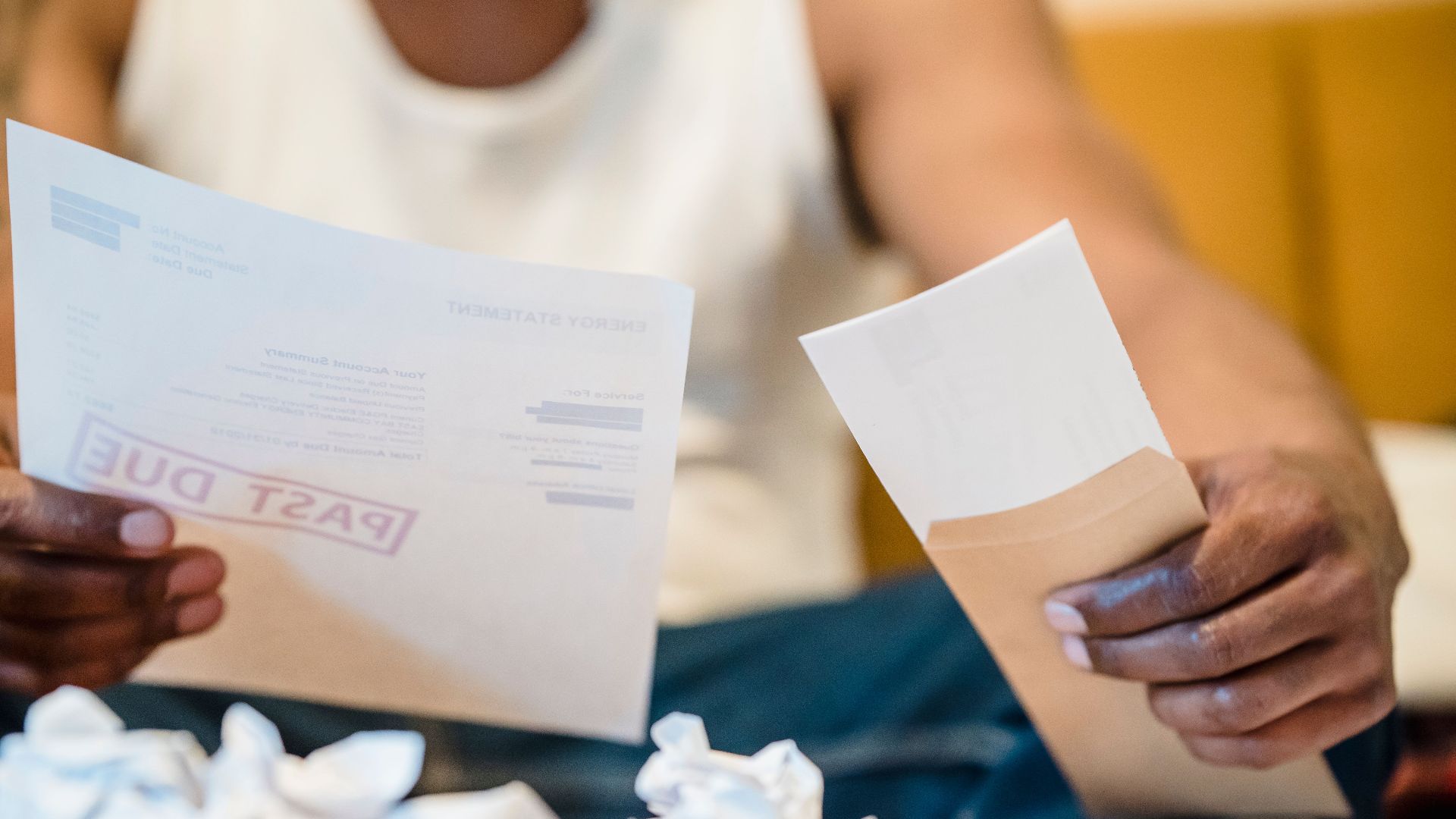 Man sitting indoors reviewing past due bills with crumpled papers on a coffee table.