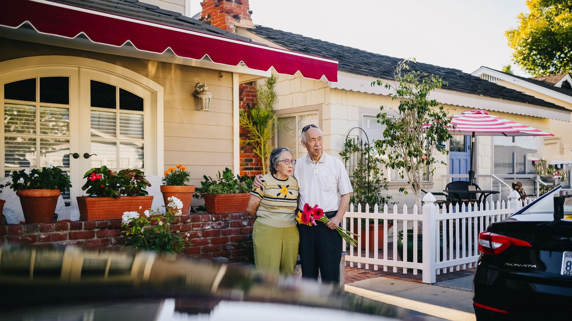 An elderly couple stands in front of a charming house holding a bouquet, capturing a serene lifestyle moment.