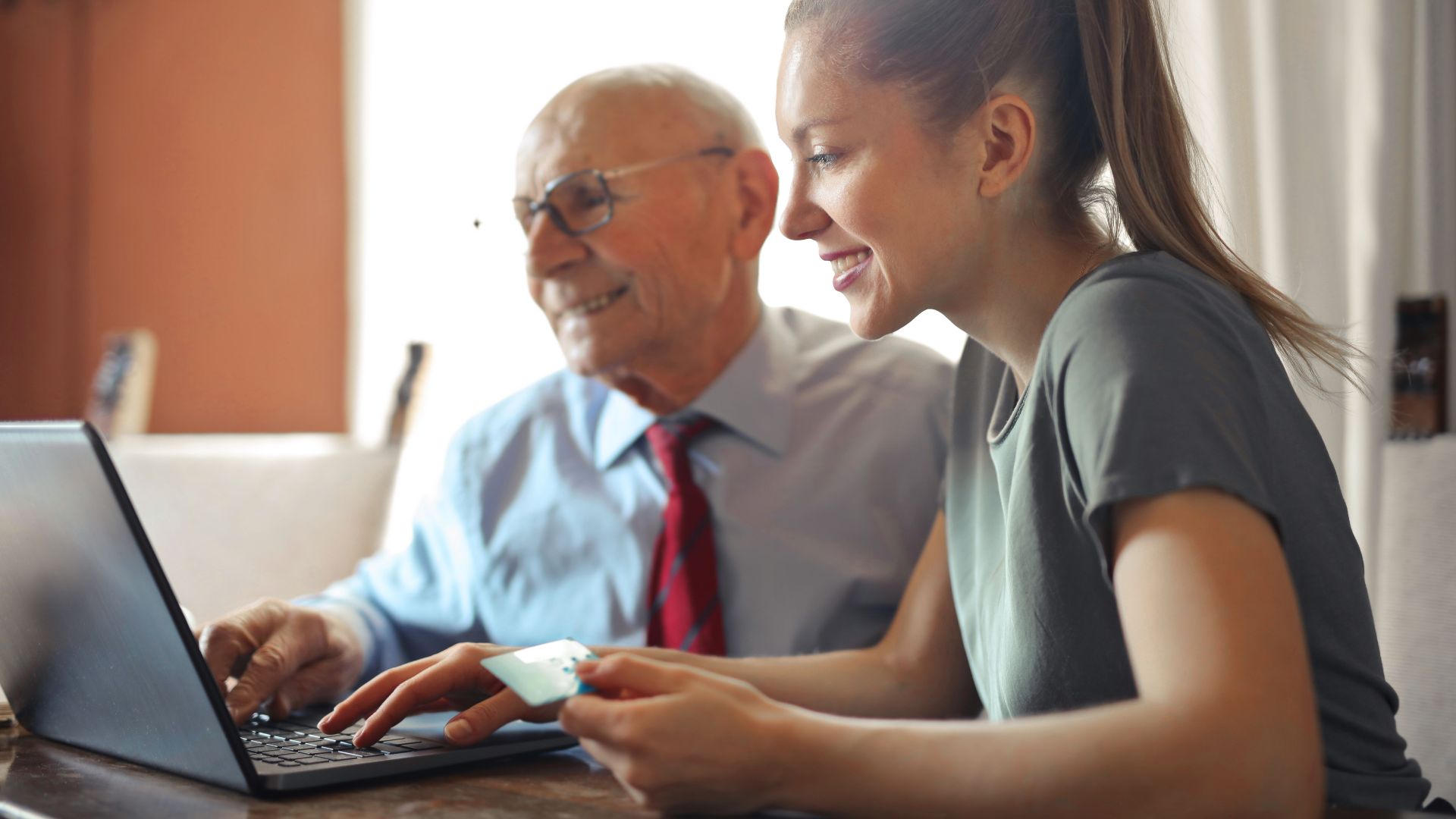 Young woman in casual clothes helping senior man in formal shirt with paying credit card in Internet using laptop while sitting at table