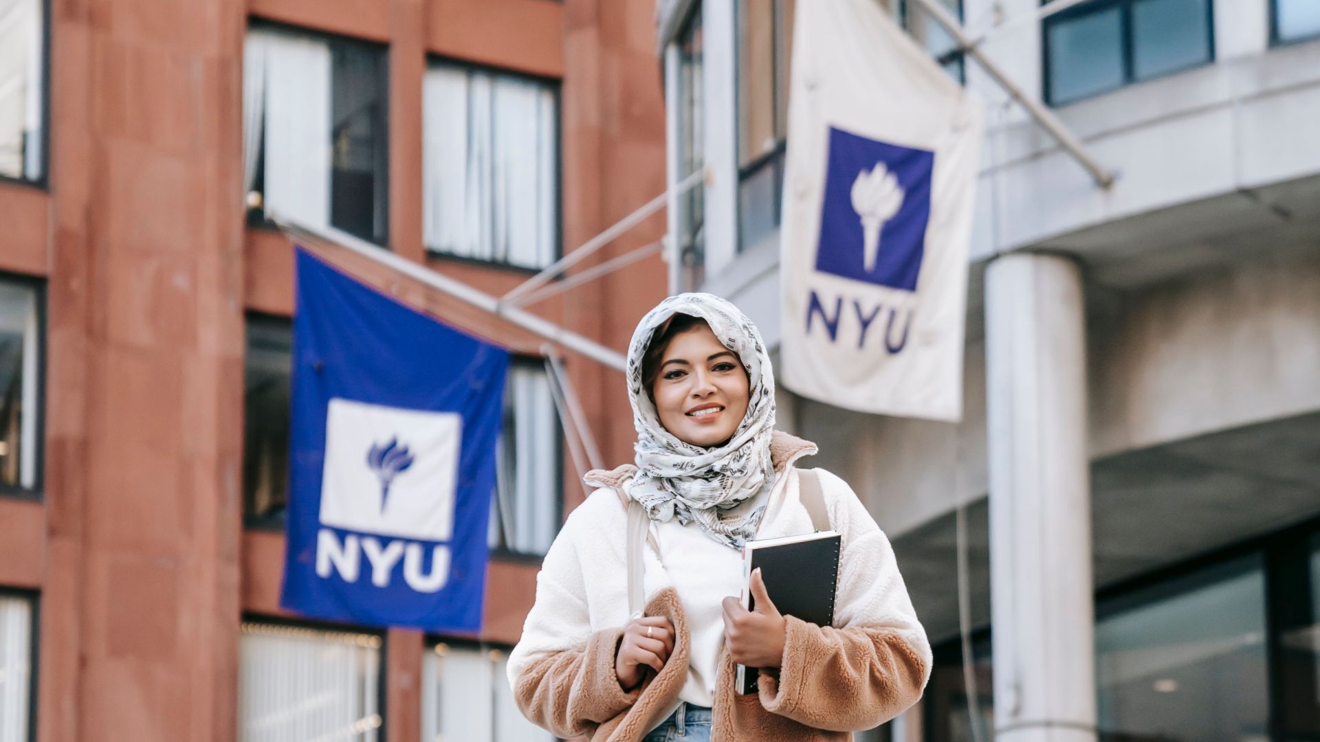 Full length of cheerful Muslim female student in stylish clothes and headscarf standing with notebook against buildings of New York University Stern School of Business in daytime